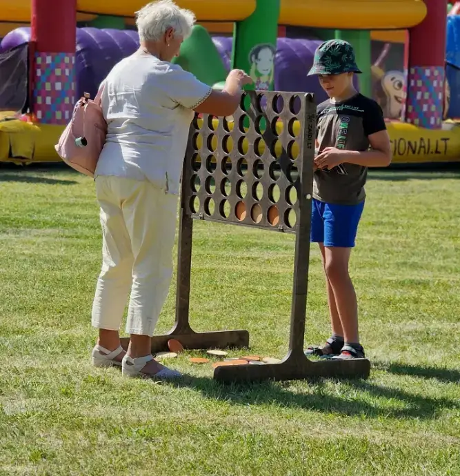 Vaikai ir suaugusieji žaidžia „Ketvirtas eilėje“ – medinį žaidimą lauke, vasaros renginyje.