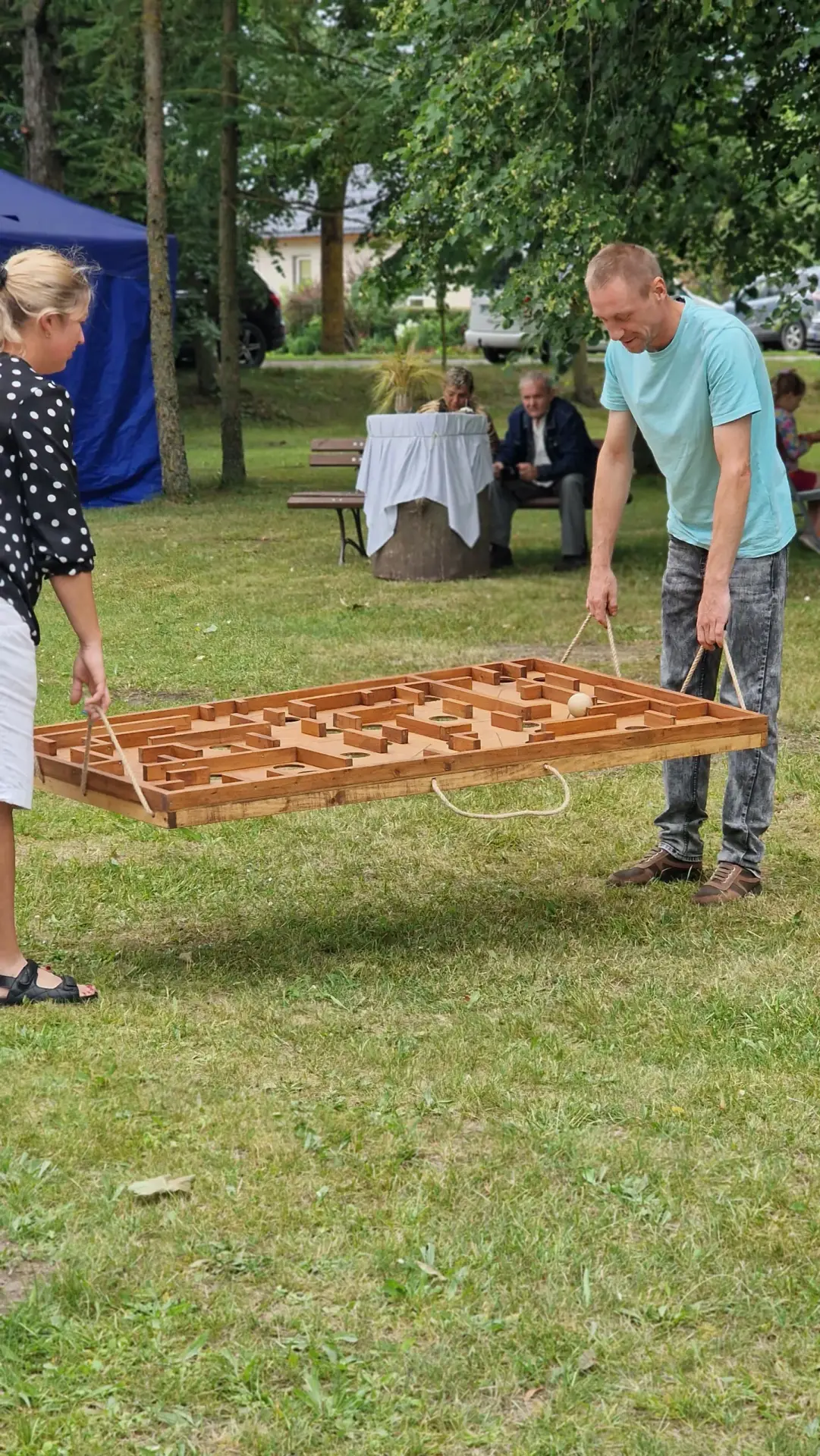 Du žaidėjai laiko medinį lauko labirintą už virvučių ir veda kamuoliuką per labirintą – horizontalus labirintas Medžio Karma