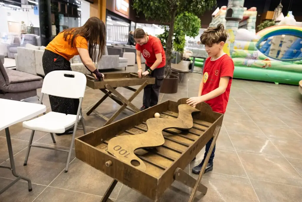 Medžio Karma medinis stalo futbolas – žaidėjos rankos valdo medinius žaidėjus ant strypo, Maltiečių sriubos renginys Šiauliuose