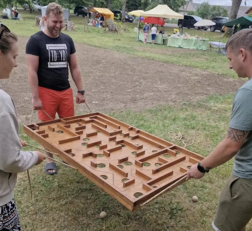 Trys žaidėjai kartu valdo medinį horizontalų labirintą lauko renginyje – komandinis žaidimas šventėje
