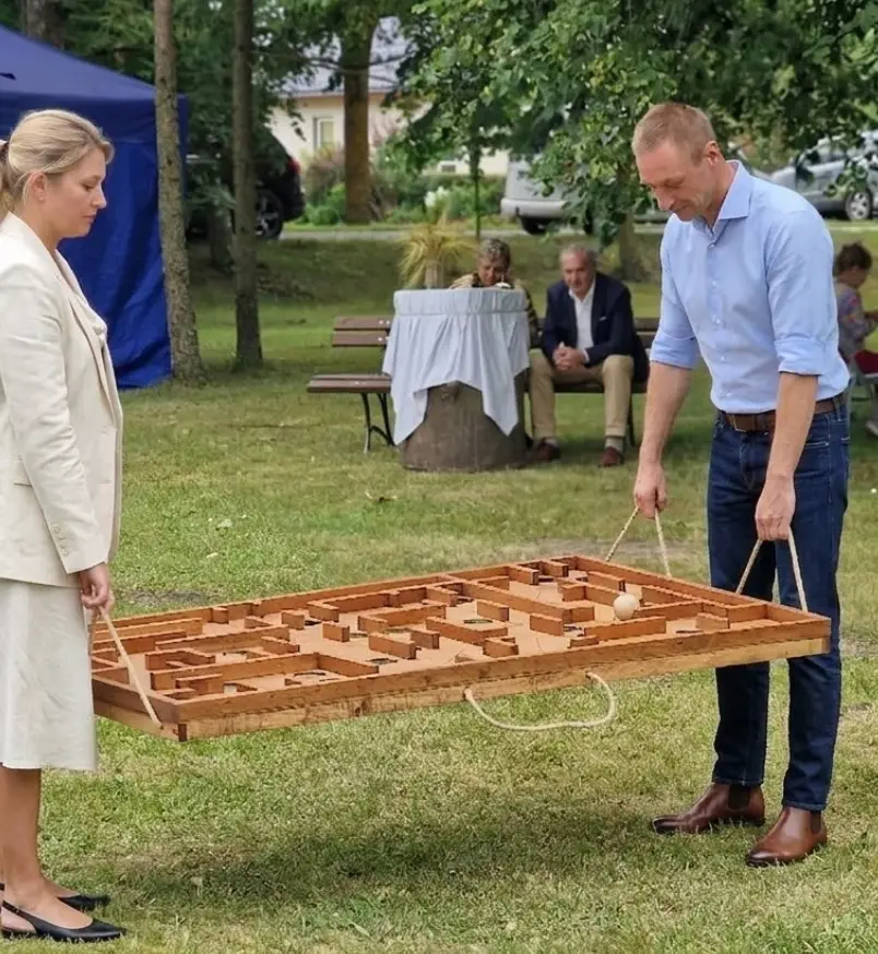 Du žaidėjai laiko medinį lauko labirintą už virvučių ir veda kamuoliuką per labirintą – horizontalus labirintas Medžio Karma