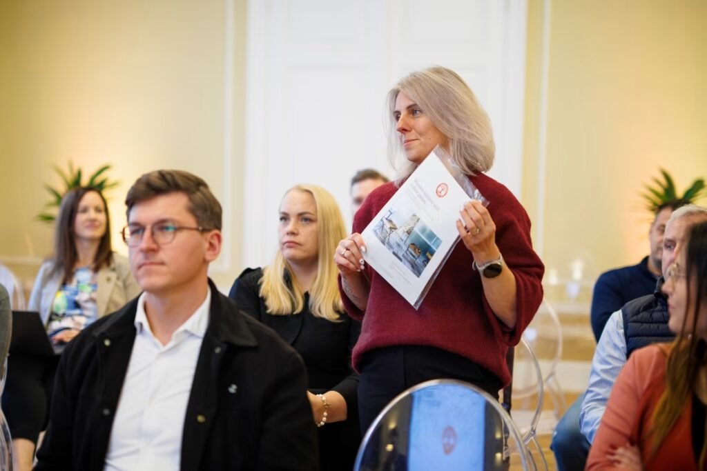 Dovilė rodo Medžio Karma medinių lauko žaidimų katalogą anglų kalba Latvijos rūmuose